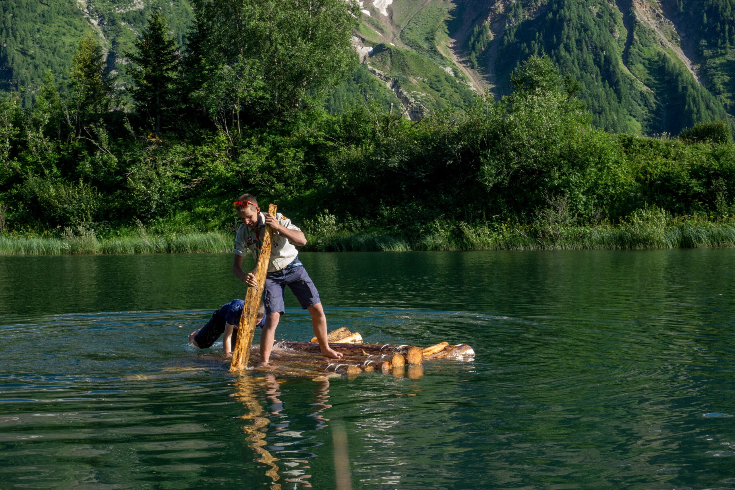 Dieses Bild zeigt zwei Pfadfinder, die mit einem Floss über einen Bergsee fahren.