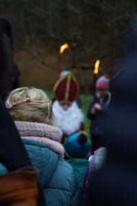 Das Bild zeigt Kinder, die Fakeln in ihren Händen halten und um Sankt Nikolaus geschart sind. Die Szenerie findet in abendlicher Dämmerung statt.