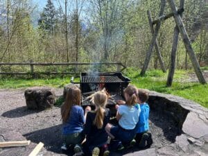 Das Bild zeigt ein paar kleine Pfadfinder, die gemeinsam Würste über einer Feuerstelle im Wald braten.
