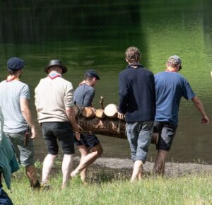 Das Bild zeigt eine Gruppe Pfadfinder, die gemeinsam ein selbstgebautes Holzfloss zum Wasser tragen.