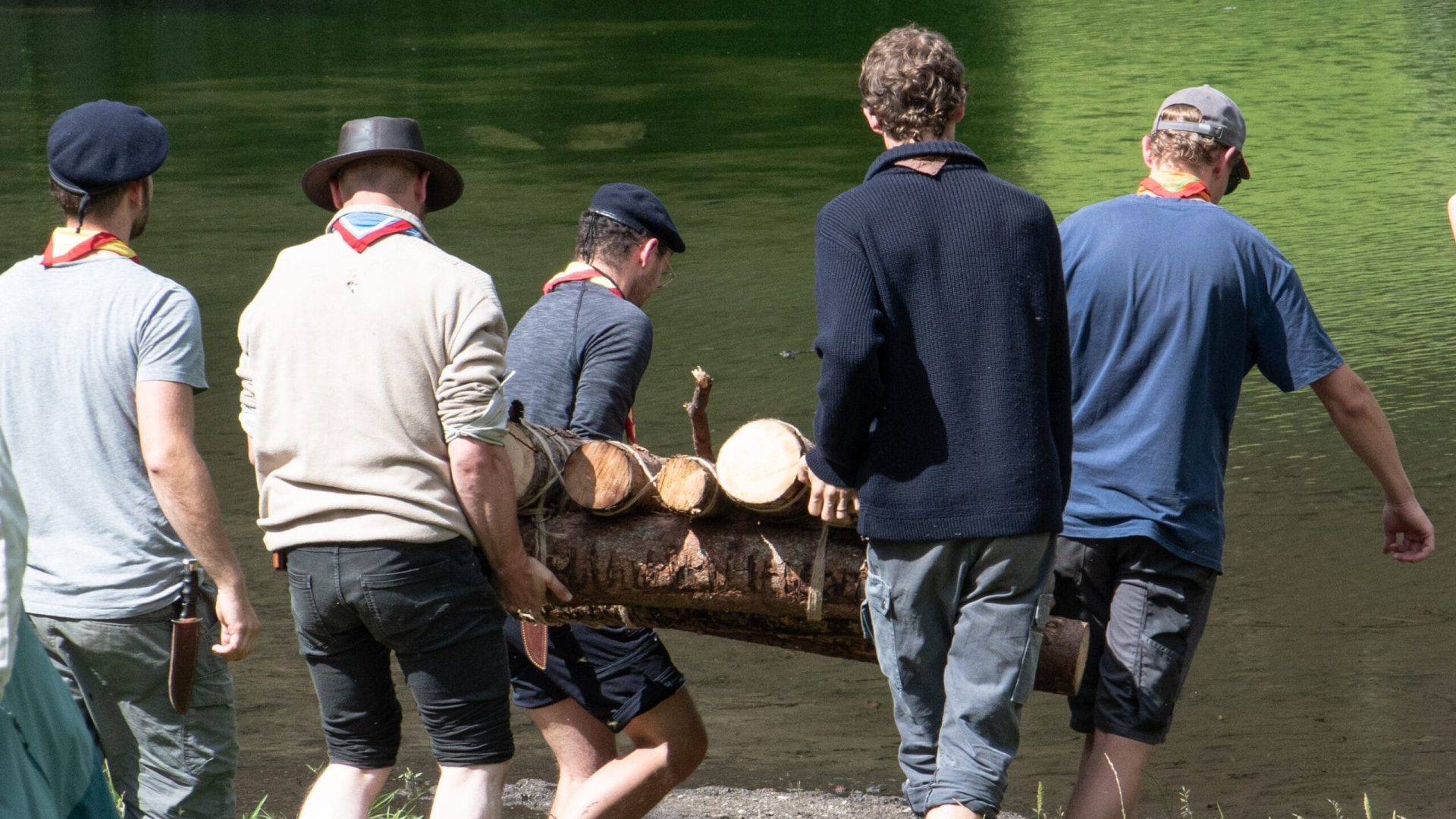 Das Bild zeigt eine Gruppe Pfadfinder, die gemeinsam ein selbstgebautes Holzfloss zum Wasser tragen.